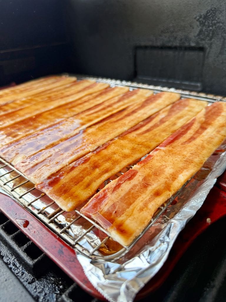 Slices of candied bacon arranged neatly on a wire rack inside a smoker, with foil underneath to catch drippings. The bacon is uncooked and glistening with a layer of seasoning.