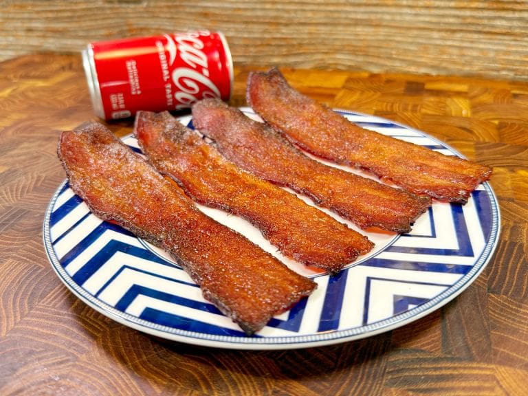 Four strips of crispy, glazed bacon on a blue-and-white zigzag patterned plate, with a partially opened can of Coca-Cola in the background, set on a wooden surface.