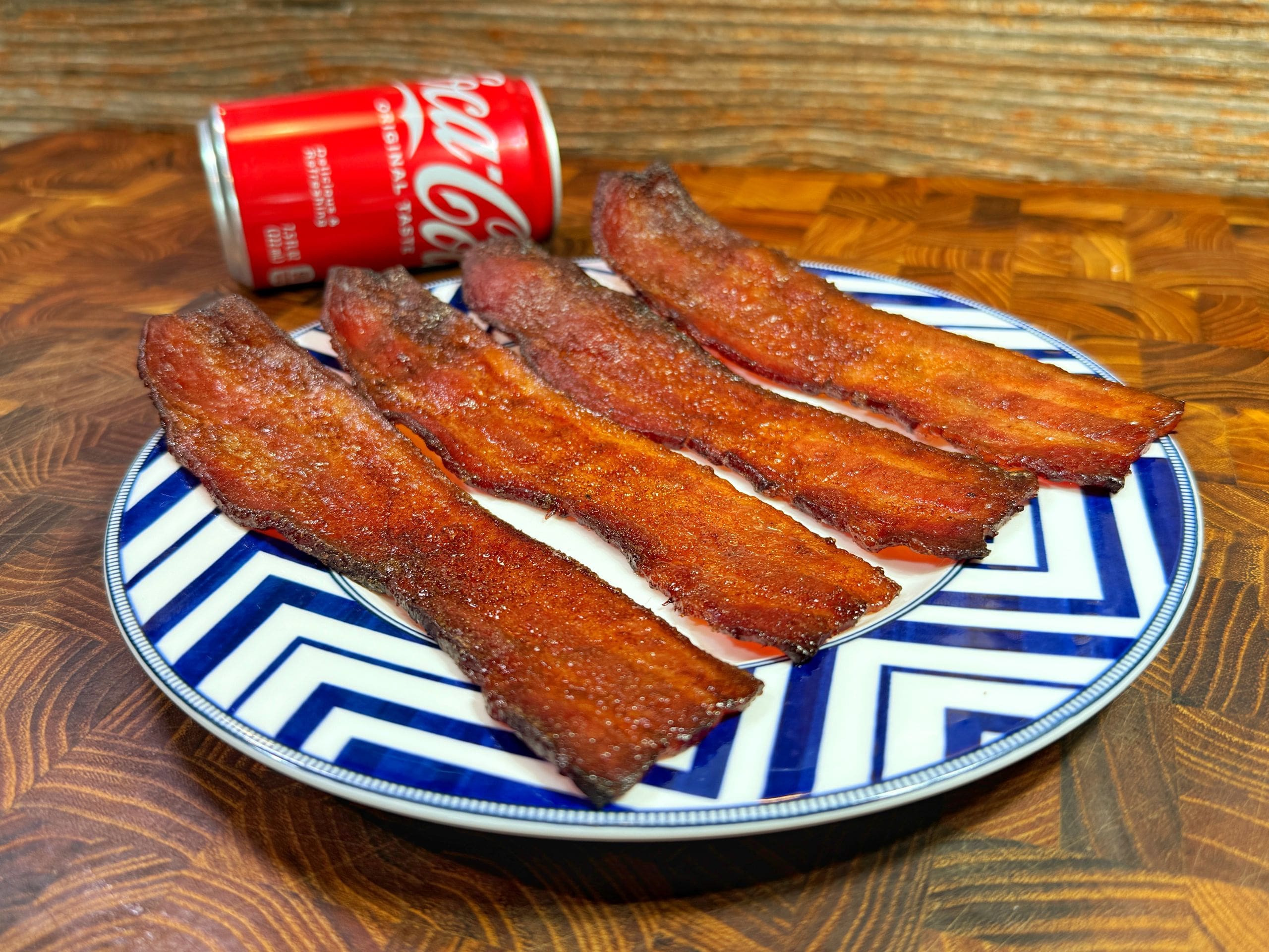 A plate with four slices of crispy bacon is placed on a patterned plate with a can of Coca-Cola in the background. The setting is on a wooden surface.