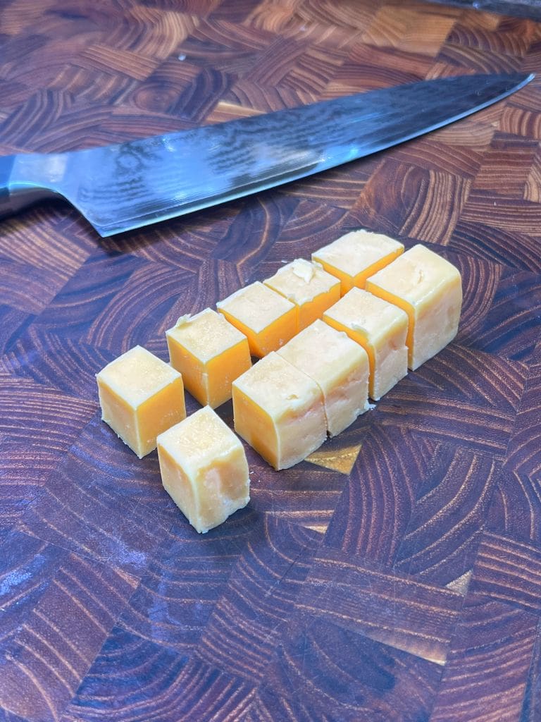 Cubes of cheddar cheese are arranged on a dark wooden cutting board, with a large stainless steel knife resting in the background.