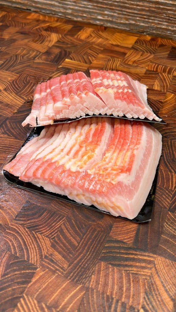 Two packs of raw bacon slices are neatly arranged on a textured wooden cutting board. The bacon features layers of pink meat and white fat, providing a striped appearance.