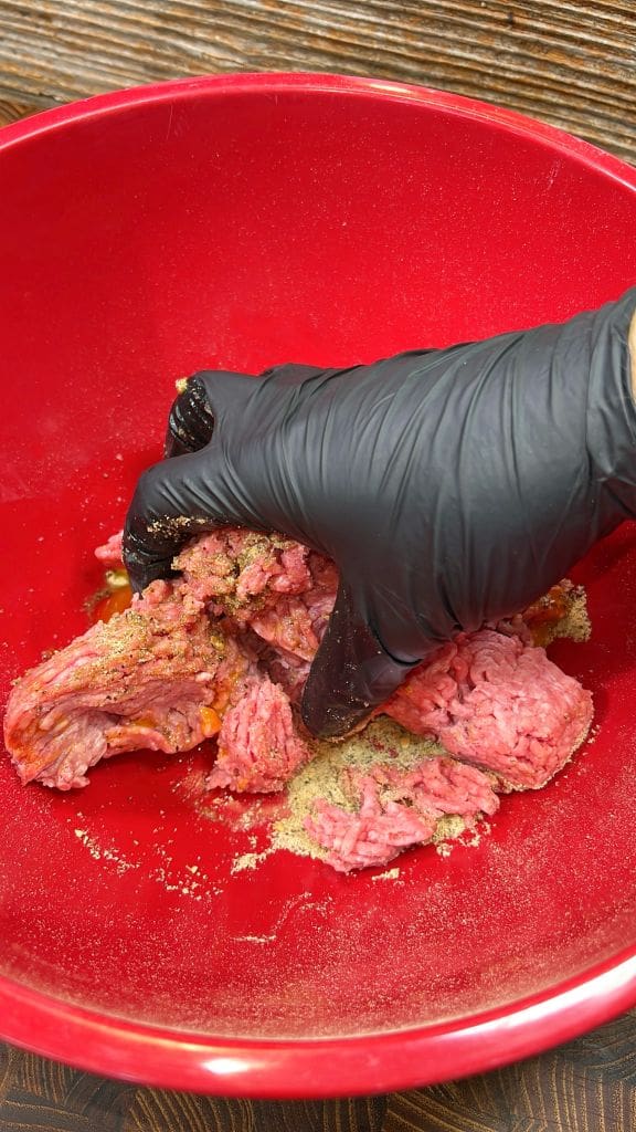 A hand wearing a black glove is mixing raw ground meat with spices in a red bowl.