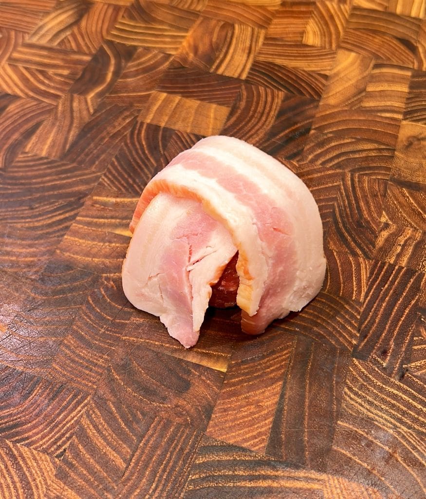 A piece of bacon-wrapped food item, possibly a date or appetizer, is placed on a wooden cutting board with a patterned surface.