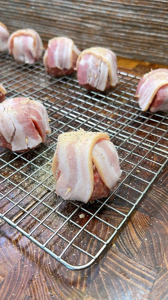 Meatballs wrapped in bacon strips, seasoned and arranged on a wire rack, ready for cooking. The wooden surface adds a rustic touch.