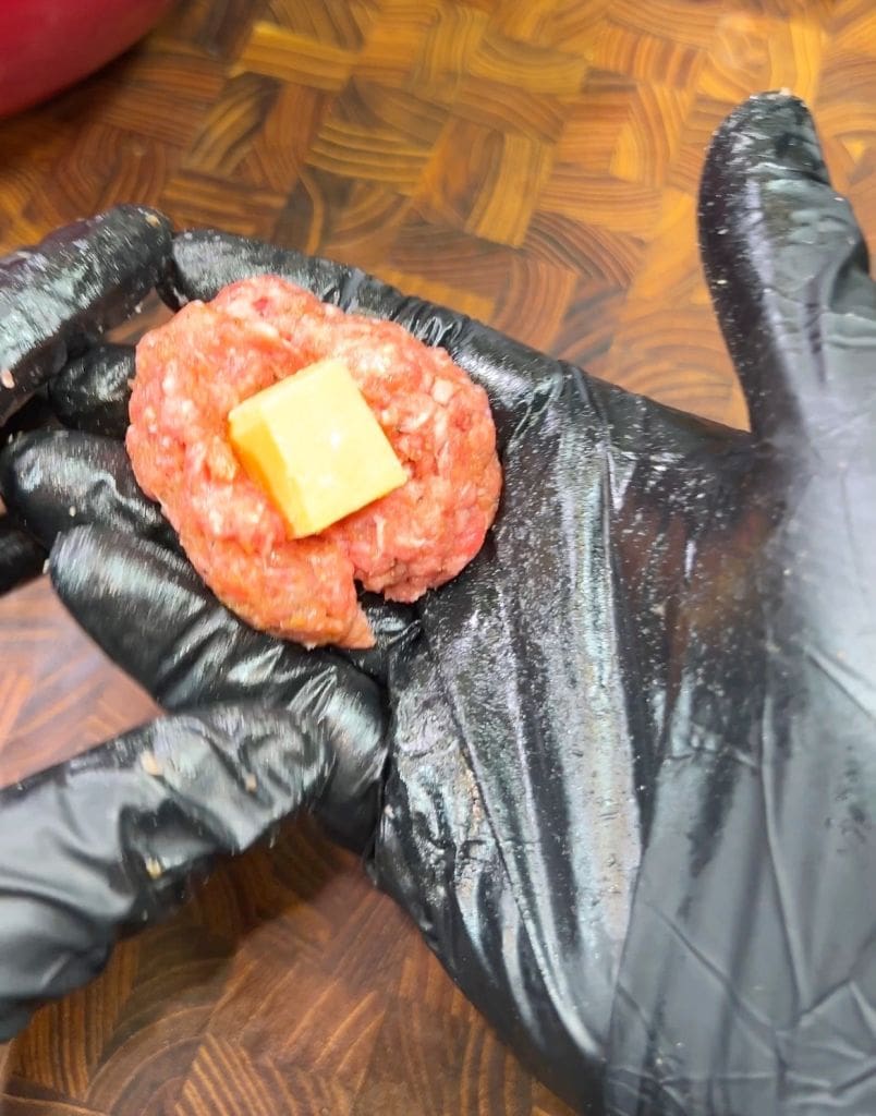 A gloved hand holding a meatball with a piece of cheese inside, on a wooden surface.