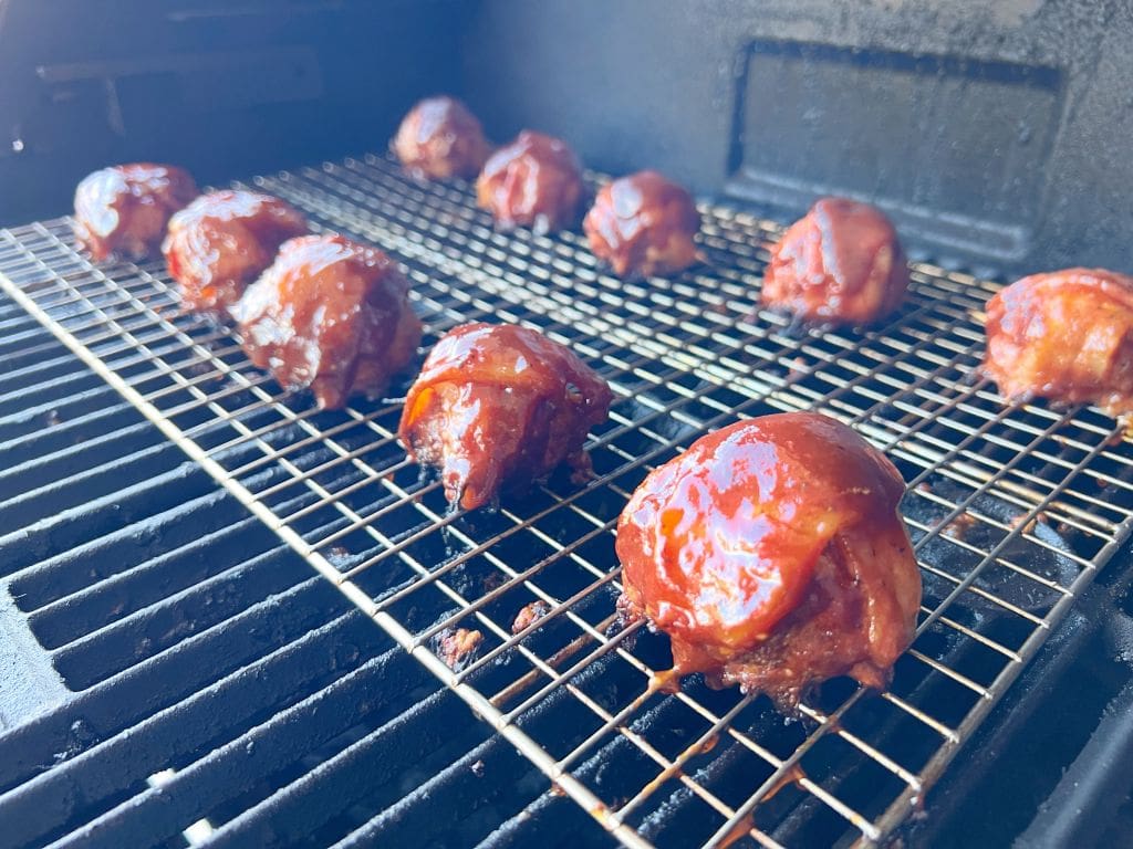 Bacon cheeseburger bombs coated in a glaze are lined up on a wire rack inside a grill, ready to be cooked. The shiny glaze is visible on each piece, and the grill grates are slightly out of focus in the background.