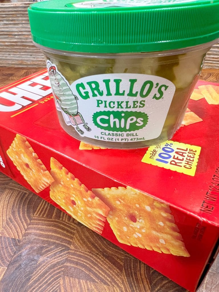 A container of Grillos Pickles Classic Dill Chips with a green lid is placed on top of a red box of Cheez-It crackers. The pickles and crackers are on a wooden surface.