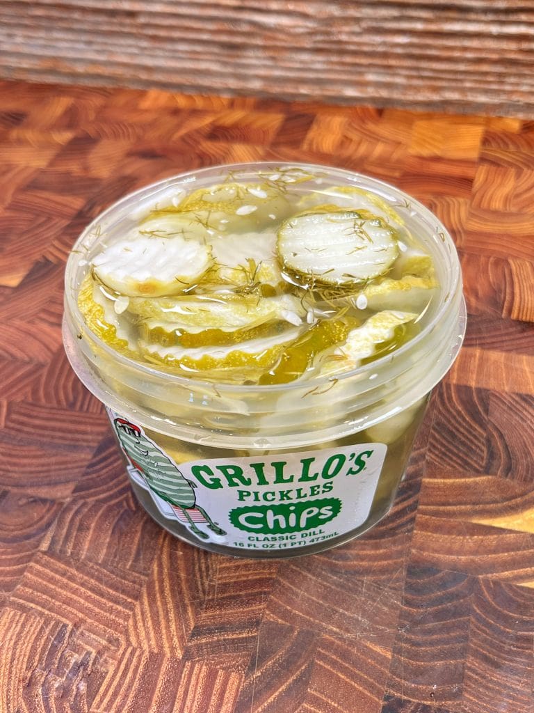 A container of Grillos Pickles rests on a wooden surface. It is filled with sliced classic dill pickles in brine. The label on the container features a pickle character and the word chips.