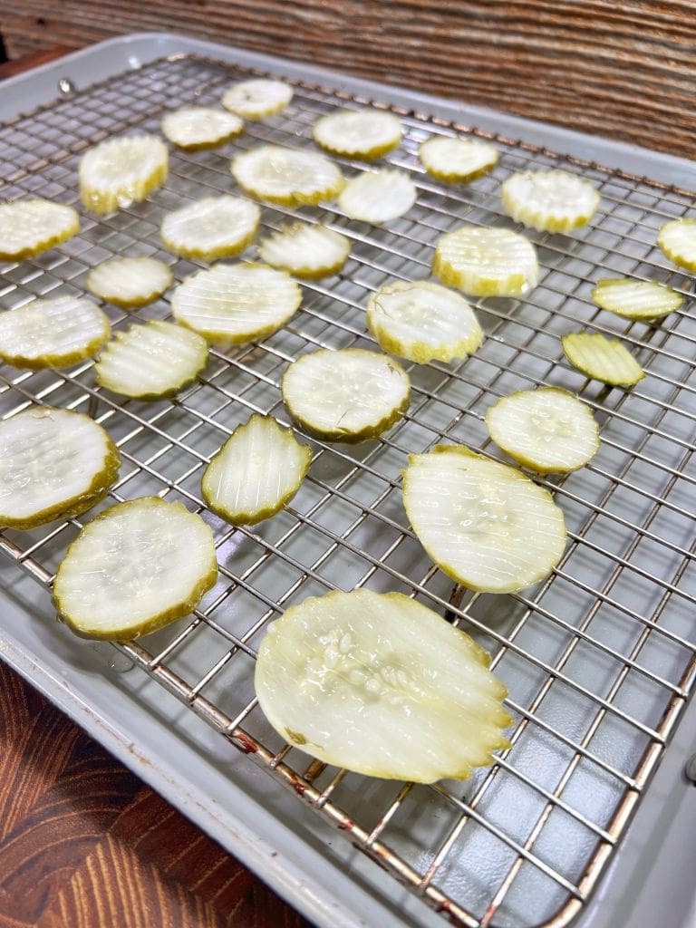 Sliced pickles arranged on a wire rack over a baking sheet. The pickles vary in size and are laid out evenly. The background features a wooden texture, adding a rustic touch to the scene.