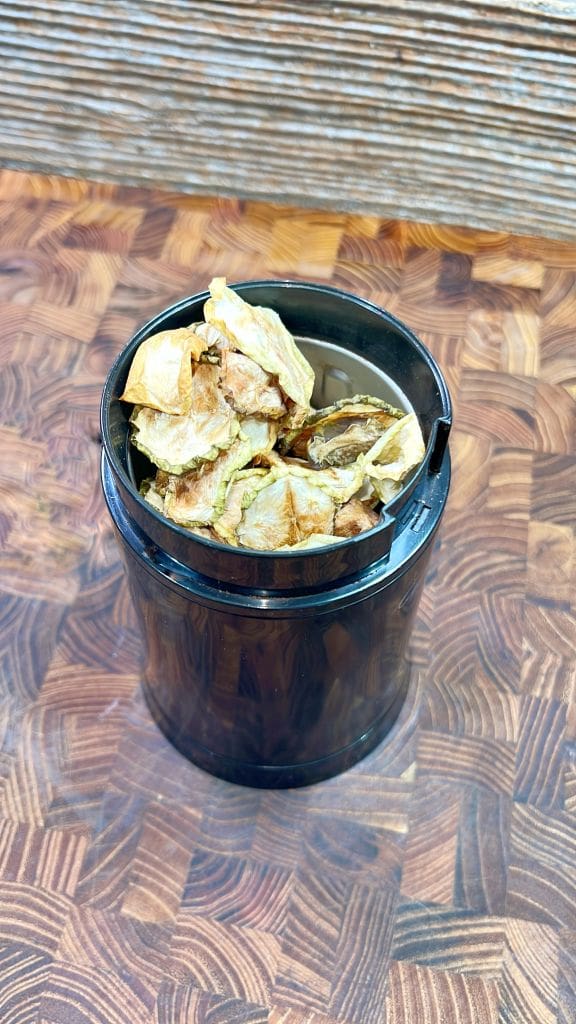 A black container filled with dried artichoke leaves is placed on a patterned wooden surface. The leaves have a brownish hue, indicating they have been dehydrated.