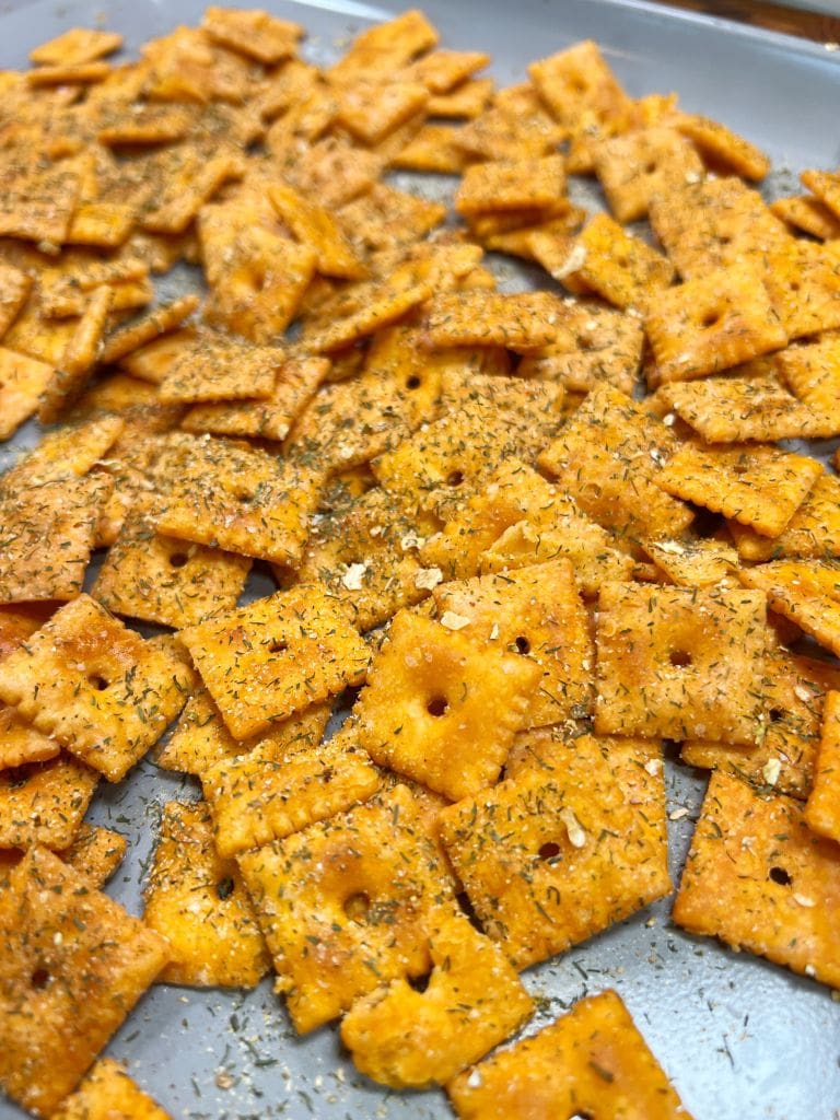 A tray filled with seasoned square cheese crackers. The crackers are sprinkled with herbs and seasoning, giving them a textured appearance.