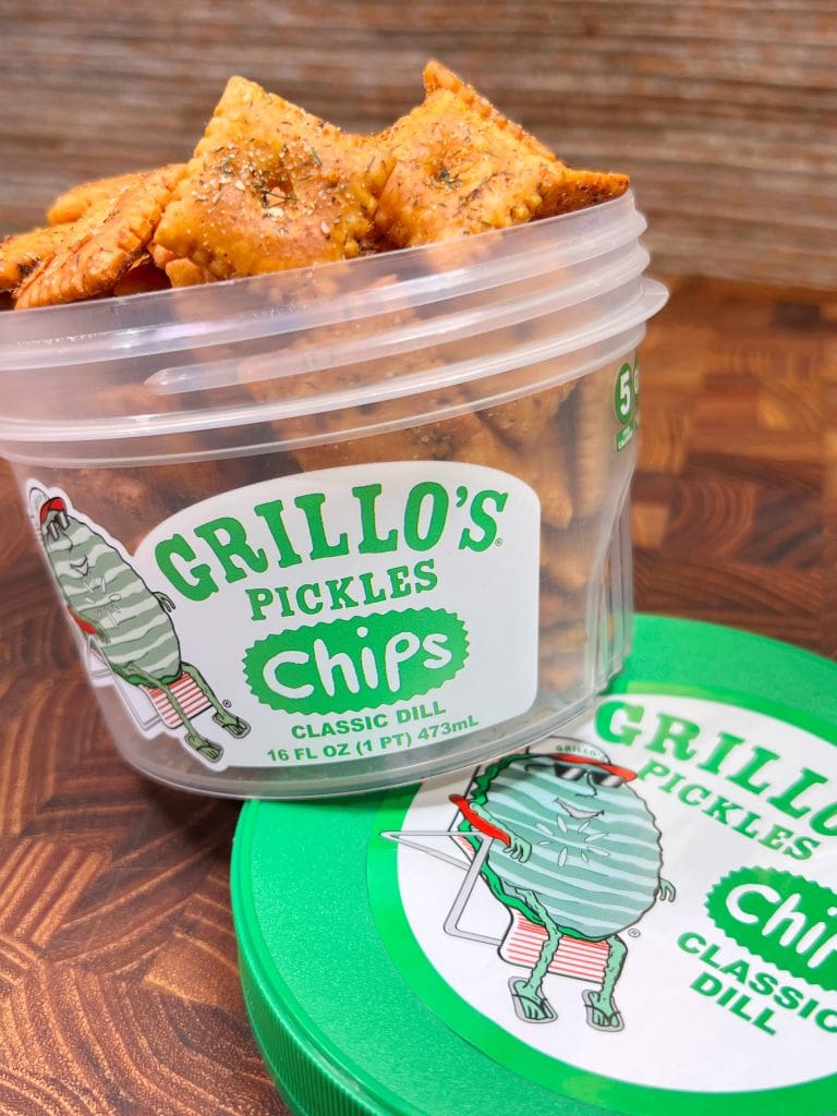 A container of Grillos Pickles Classic Dill Pickle Cheez its  is open, showing seasoned square chips inside. The lid is lying nearby. Both the container and lid feature a pickle character wearing sneakers. The background is a wooden surface.
