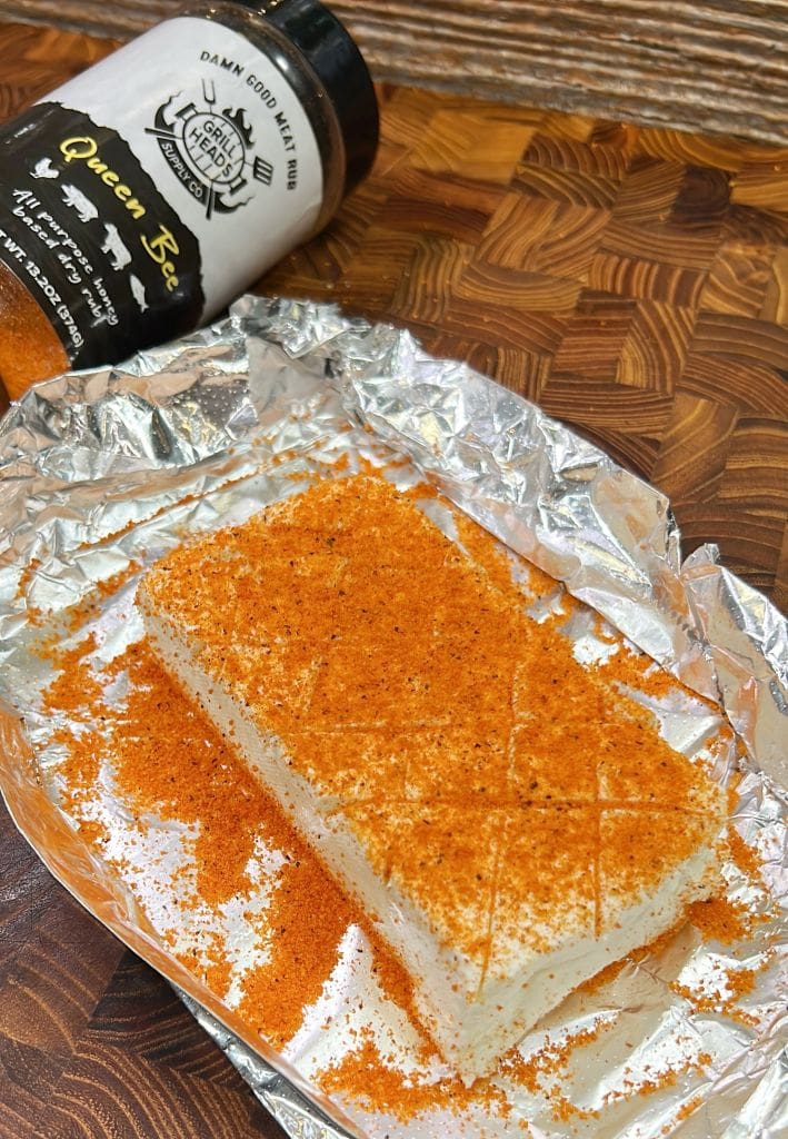 A rectangular block of cheese covered with a spicy seasoning is placed on aluminum foil. A bottle labeled “Queen Bee” seasoning is lying next to it on a wooden surface. The cheese has a crosshatch pattern on its top.