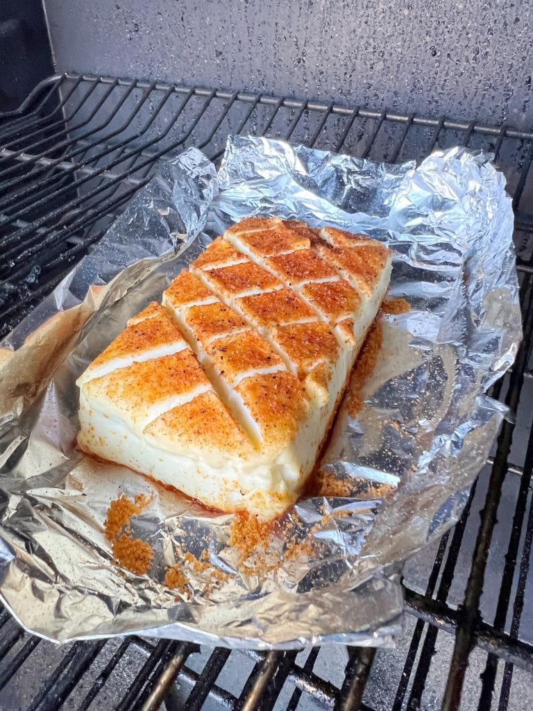 A large block of cheese with a crisscross pattern on top is placed on foil inside a grill. The cheese is seasoned with a reddish spice mix and appears slightly melted around the edges.