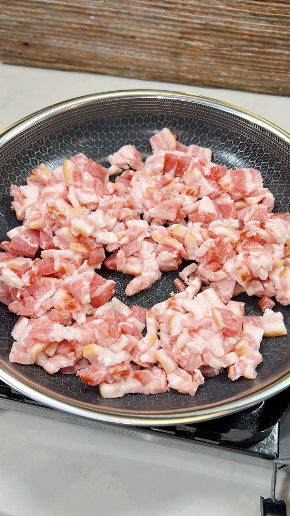 Chopped bacon pieces frying in a pan on a stovetop.