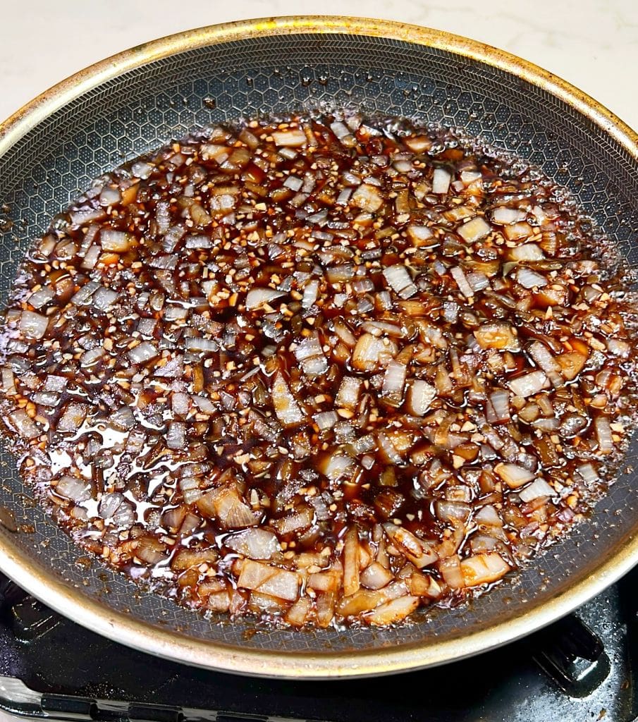 A close-up of a frying pan with chopped onions caramelizing in a dark sauce on a stovetop. The onions are evenly spread, glistening as they cook in the rich sauce.