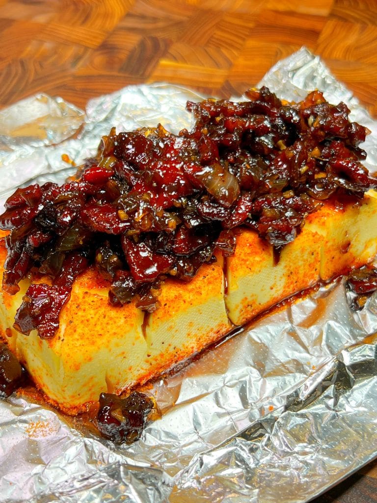 A block of tofu topped with a savory mixture, likely caramelized onions and spices, sitting on aluminum foil. The tofu is lightly seasoned with paprika or a similar red spice, and the background has a wooden texture.