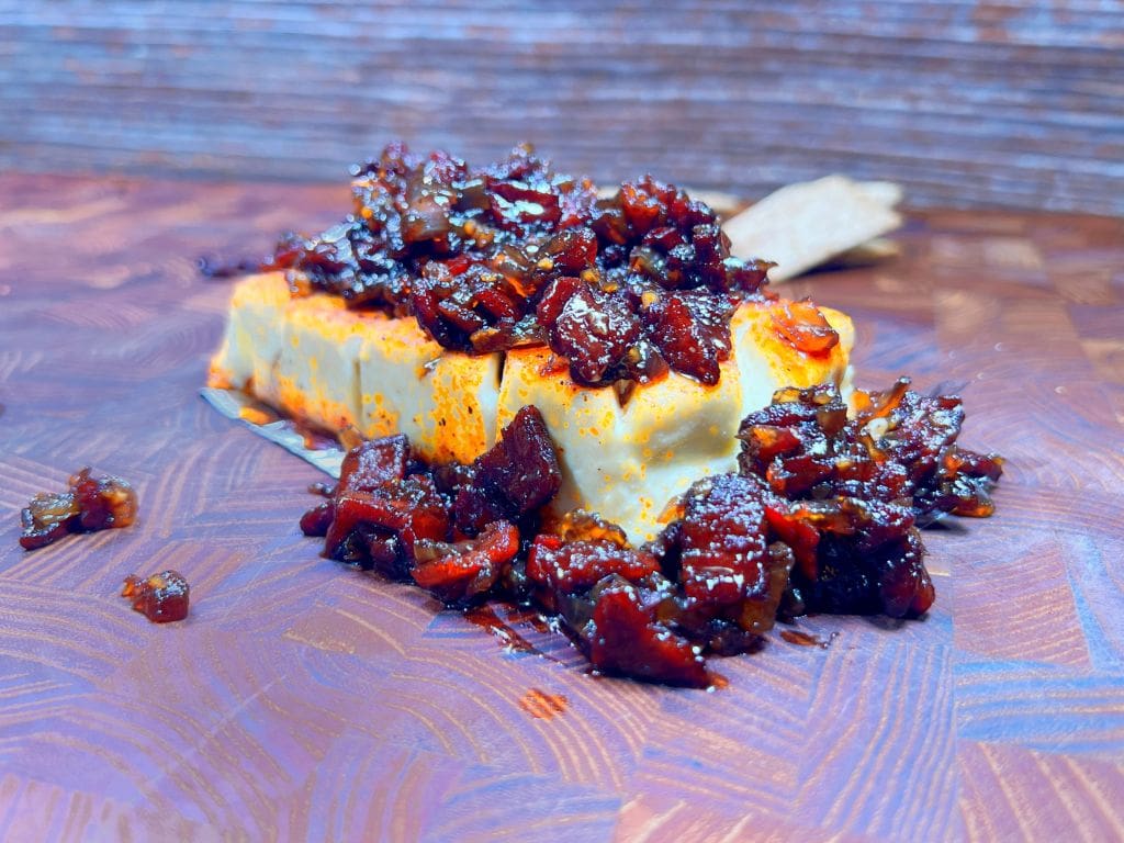 A block of smoked cream cheese is topped with a generous amount of spicy, caramelized bacon jam.  It sits on a textured wooden surface, with a cracker partially visible in the background, adding a rustic and appetizing appeal.