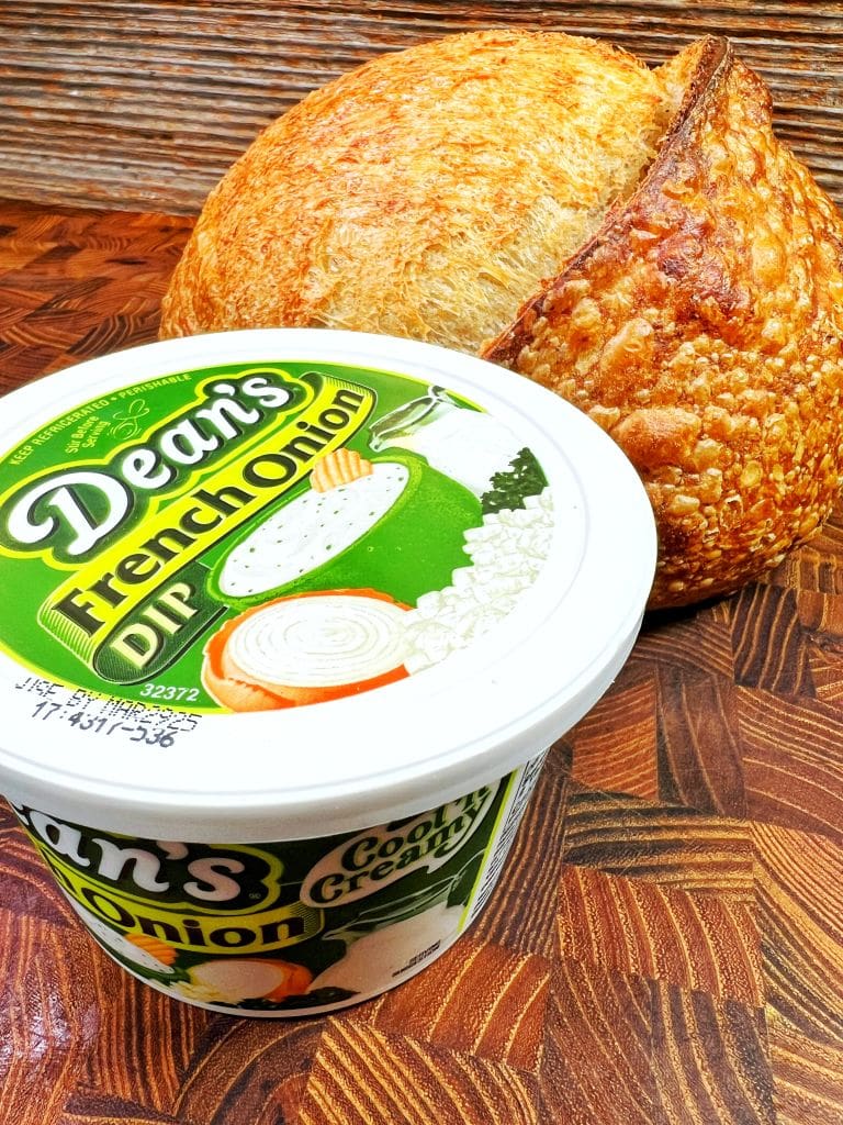 A round loaf of rustic bread is placed next to a container of Deans French Onion Dip on a wooden surface. The bread has a golden crust, and the dip container features images of onions and green accents on the label.