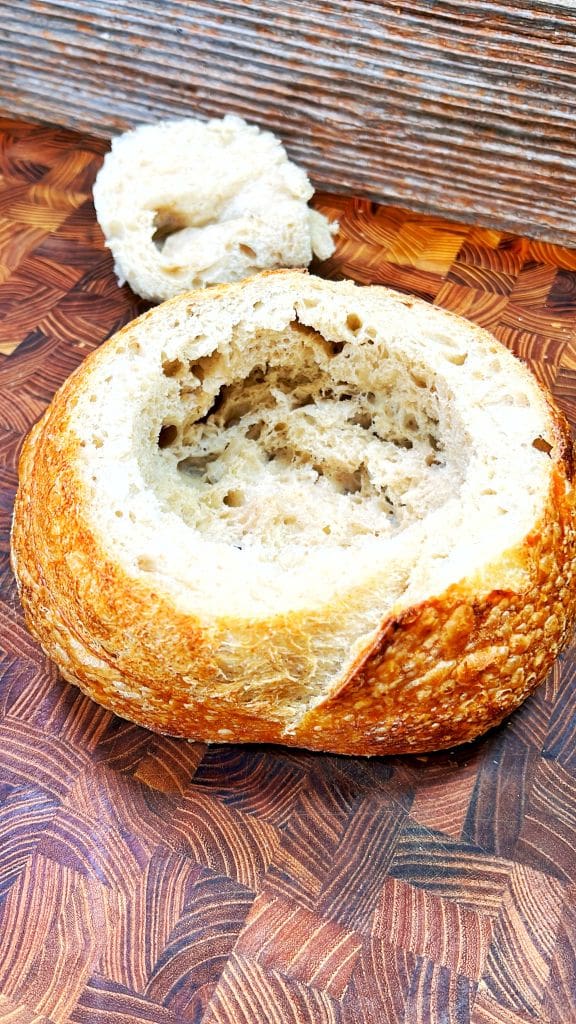 A hollowed-out round loaf of bread sits on a textured wooden surface, with a piece of the removed bread placed beside it.