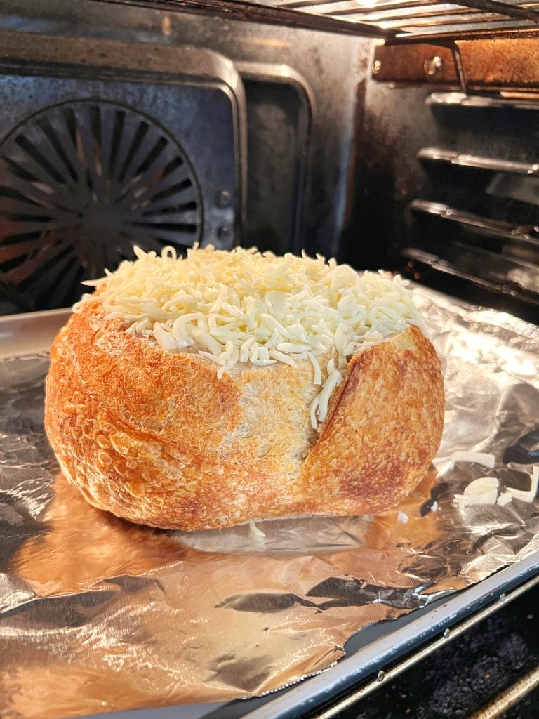 A round loaf of bread, hollowed out and filled with shredded cheese, sits on a sheet of aluminum foil inside an oven. The bread has a golden crust, and the ovens interior is dark with visible heating elements.