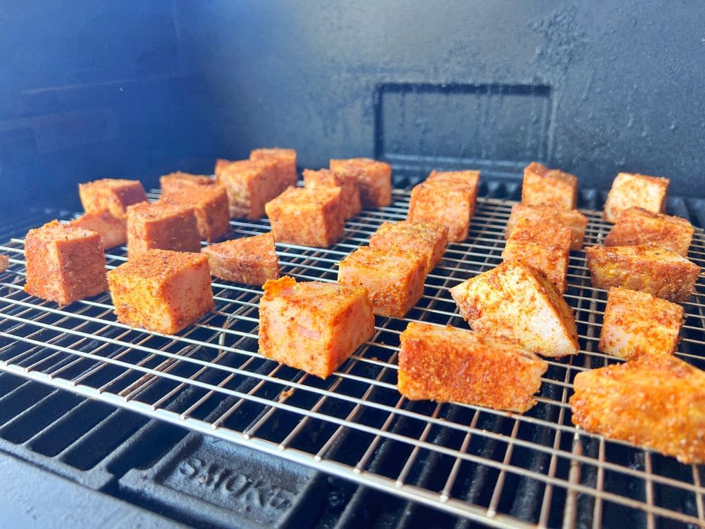 Cubes of seasoned tofu are arranged on a wire rack inside a smoker, ready to be smoked. The tofu is coated with a reddish spice mix, giving a flavorful appearance against the black interior of the smoker.