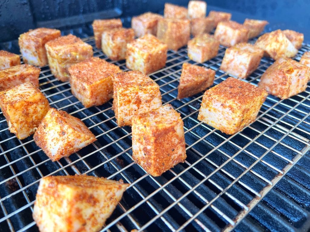 Cubed pieces of seasoned food, likely tofu or paneer, are placed on a metal grill rack. The cubes are coated with a reddish seasoning and are being cooked evenly on the grill. The background shows a dark cooking surface.