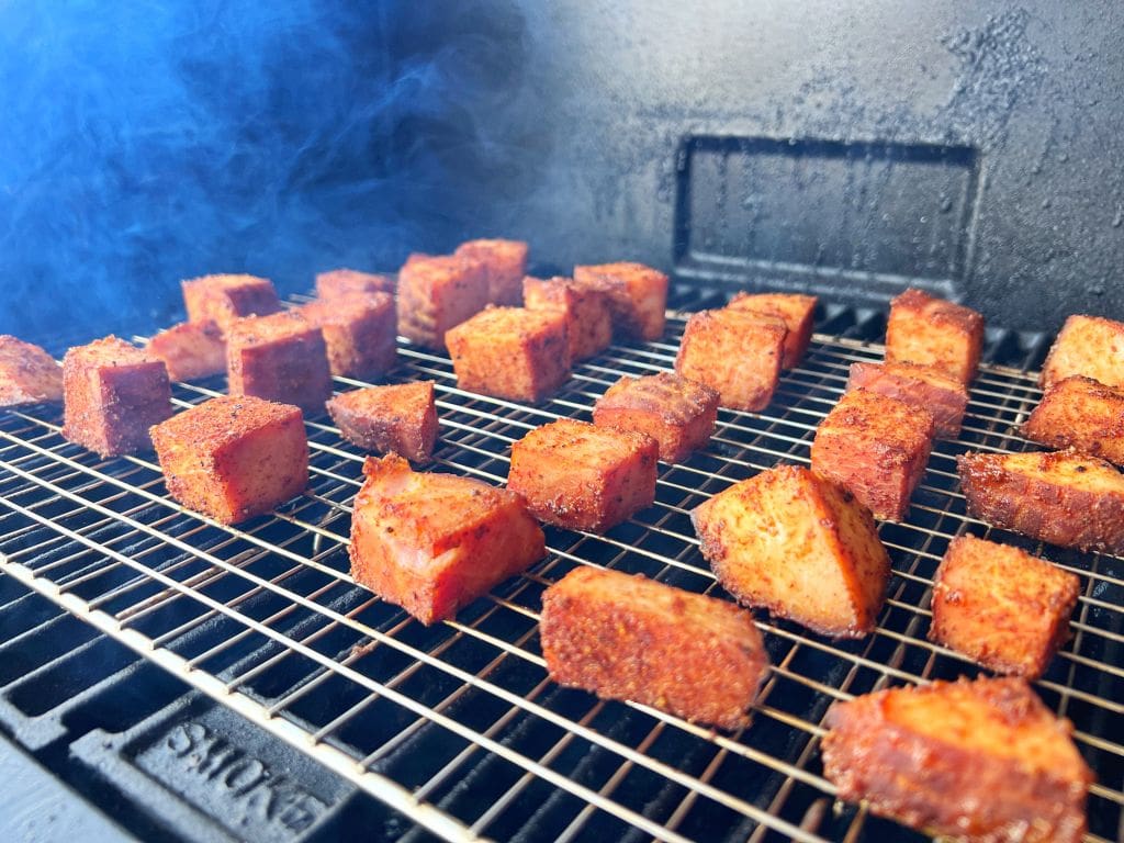 Cubes of spiced ham burnt ends arranged on a grill rack inside a smoker, with tendrils of smoke rising around them, indicating they are being cooked slowly for a smoky flavor. The meat appears to be seasoned with a reddish spice rub.