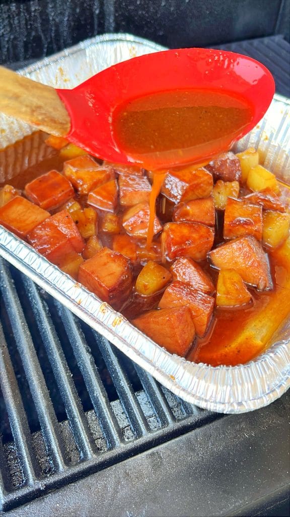 A red spoon drizzles sauce over glazed pineapple and meat chunks in a foil tray on a grill.