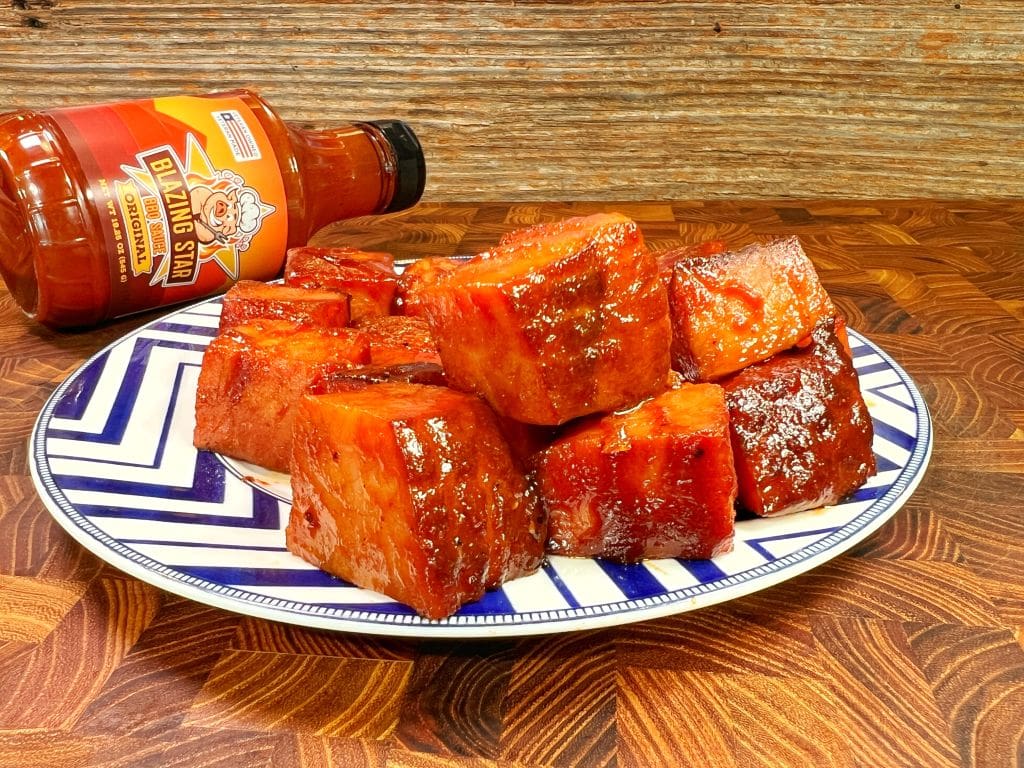 Plate of glazed pork belly cubes on a blue-and-white patterned dish, with a bottle of BBQ sauce labeled Blazing Star on its side in the background. The wooden surface adds a rustic touch.