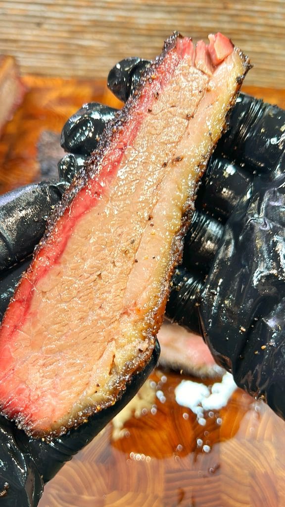 A close-up of a gloved hand holding a slice of smoked brisket. The meat has a prominent pink smoke ring and a juicy, tender texture.