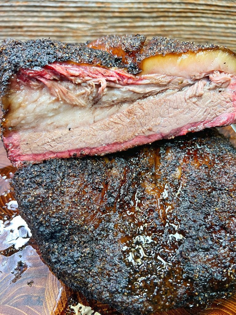 Close-up of two thickly sliced pieces of smoked brisket, showcasing a dark, seasoned crust and a moist, tender interior with a reddish smoke ring. The wooden background is lightly stained with juices from the meat.