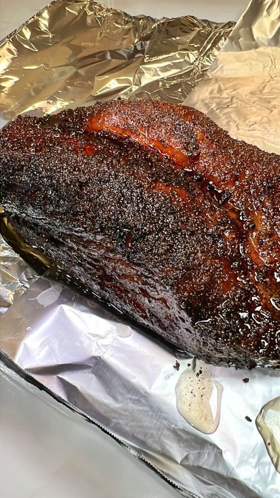 A close-up of a perfectly cooked, seasoned brisket done resting on foil. The dark, crispy crust glistens with moisture, and the foil reflects light, emphasizing the juiciness and rich texture of the meat.