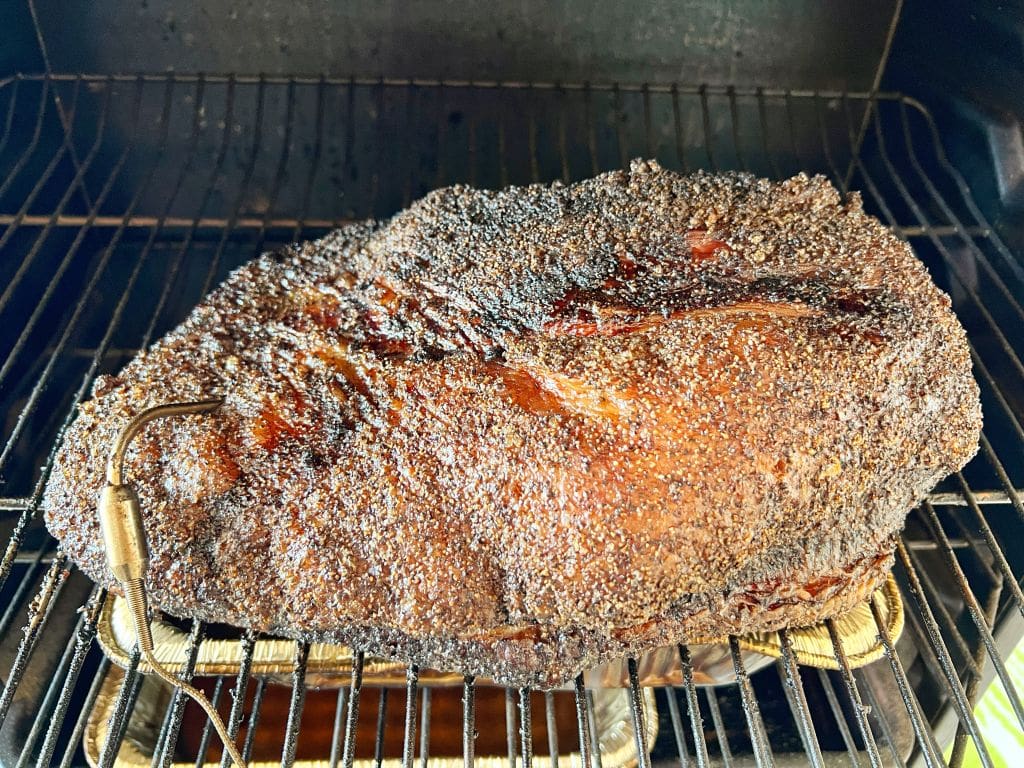 A large, perfectly smoked beef brisket sits on a metal rack inside a smoker. The meat is coated in a dark, crispy bark, showing a rich texture and savory appearance. A meat thermometer probe is inserted on the left side.