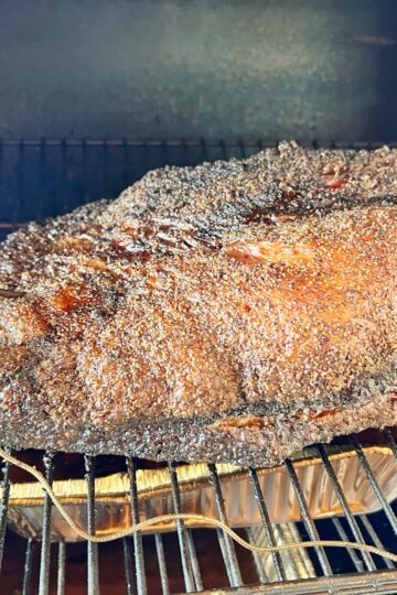 A large piece of smoked brisket with a crispy, dark crust is cooking on a grill. A meat thermometer is inserted into the brisket, and it rests on a foil tray inside the smoker.