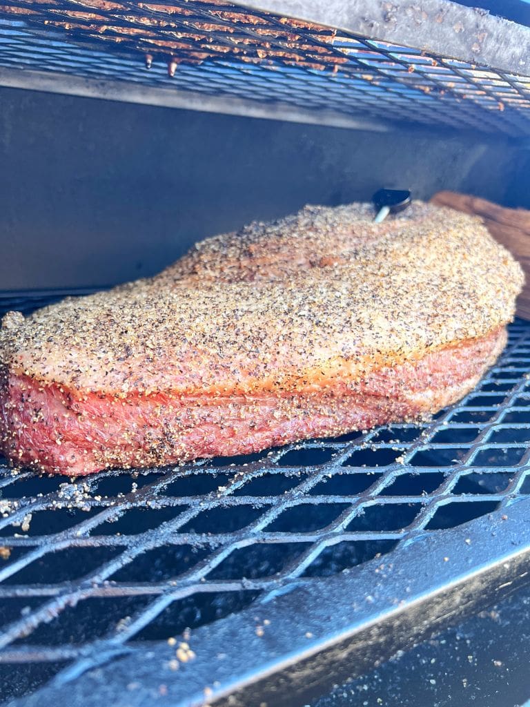 A seasoned brisket is being smoked on a grill, with a thick crust of spices visible. The grill grate holds the meat, which is cooking slowly, enveloped in smoky hues.