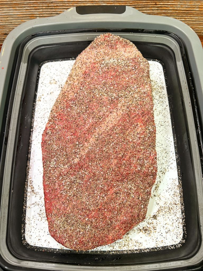 A seasoned raw meat slab on a white surface inside a black tray, sprinkled with coarse black pepper and other spices, ready for cooking.