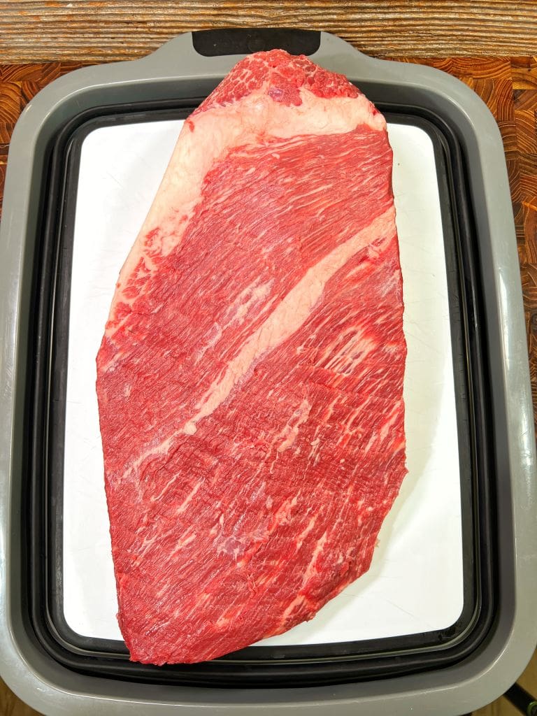 A large raw beef brisket with visible marbling sits on a white tray with a black and gray border, placed on a wooden table.