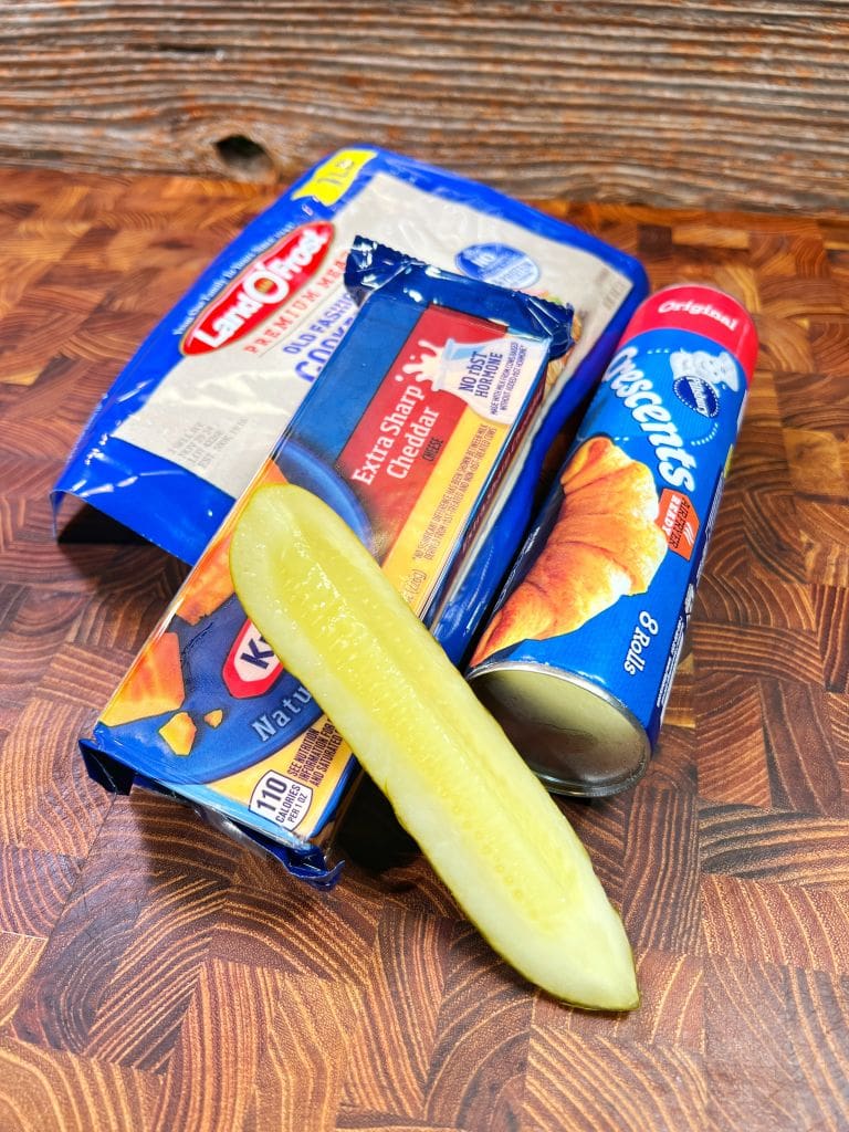 A block of cheddar cheese, a package of mozzarella cheese slices, a can of crescent rolls, and a sliced dill pickle are arranged on a wooden surface in prep for making ham and cheese rollup