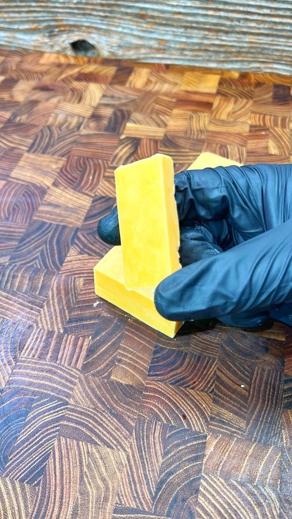A person wearing black gloves holds two stacked yellow soap bars on a textured wooden surface.
