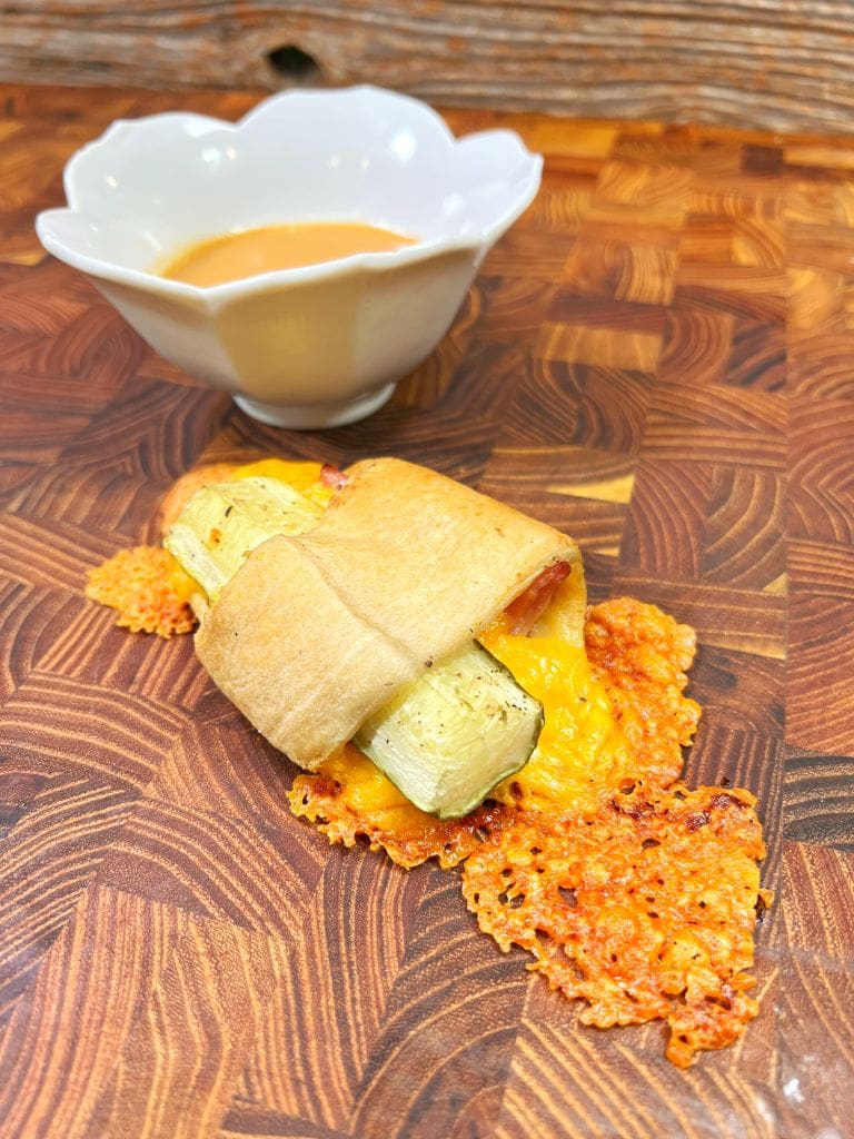 A zucchini wrapped in puff pastry with melted cheese oozing out, placed on a wooden board. In the background, there is a small white bowl containing an orange dipping sauce.