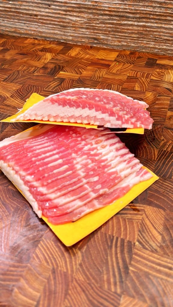 Two packages of sliced bacon are placed on a wooden countertop, their pink and white marbled appearance perfect for creating a delicious snack recipe. One package slightly overlaps the other against the detailed, checkered wood pattern, evoking thoughts of bacon-wrapped pizza rolls.