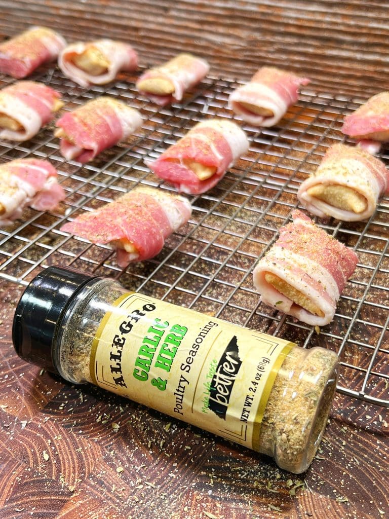 Seasoned bacon-wrapped pizza rolls rest on a wire rack, with a bottle of Allegro Garlic & Herb poultry seasoning in the foreground. These delightful bites sit on a wooden surface, ready to be cooked—a perfect snack recipe for any occasion.