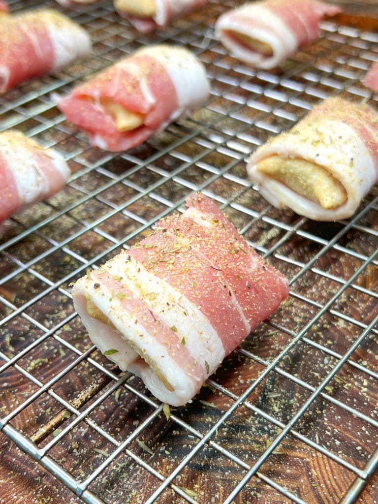 Close-up of bacon-wrapped pizza rolls, seasoned with spices. These delectable snack recipe pieces are placed on a metal rack over a wooden surface, ready for cooking or baking.