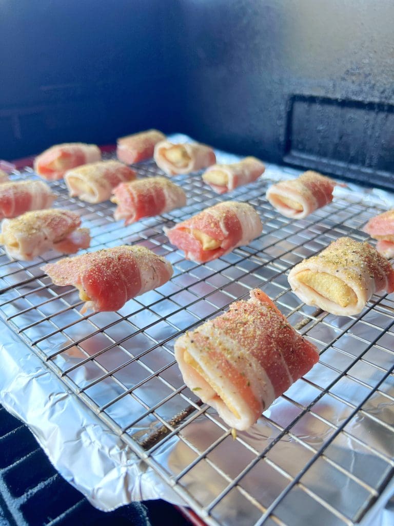 Bacon-wrapped chicken pieces, seasoned with spices, resemble delectable pizza rolls as they sit on a wire rack lined with foil, ready to be cooked into a scrumptious snack recipe.