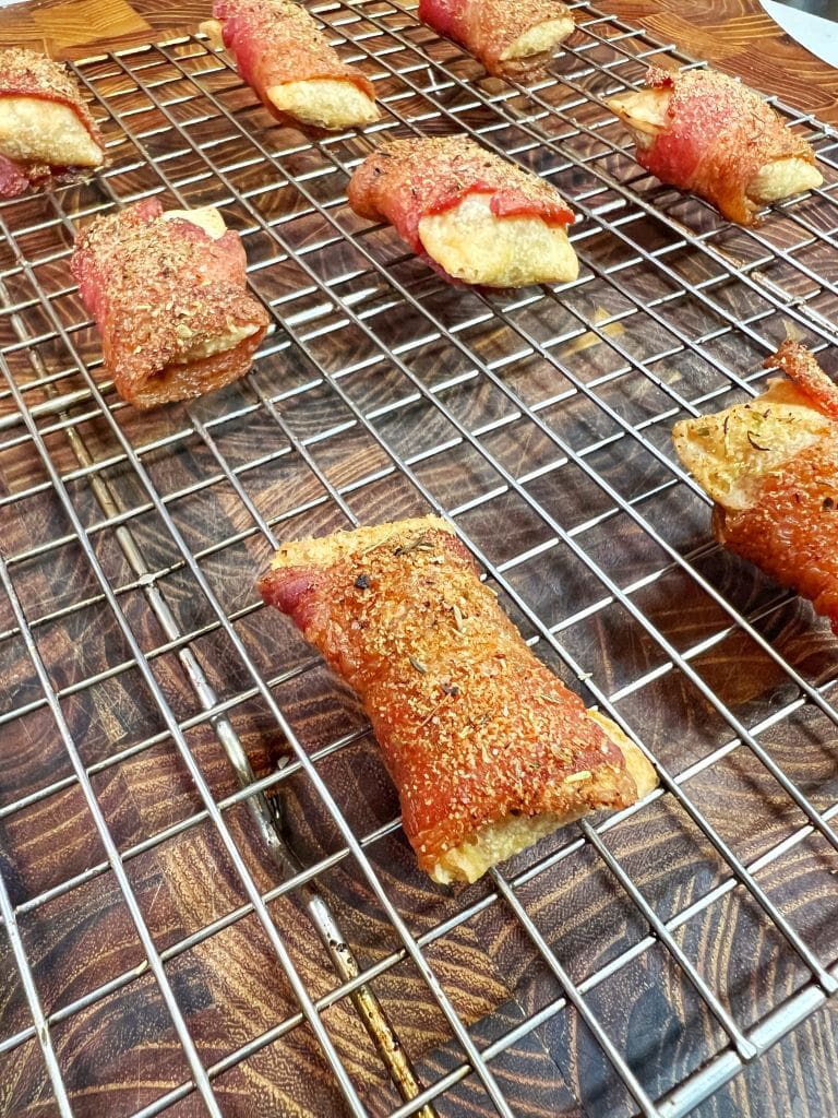 Bacon-wrapped snacks on a cooling rack, sprinkled with seasoning. They rest on a wooden surface, showcasing a crispy, golden appearance.