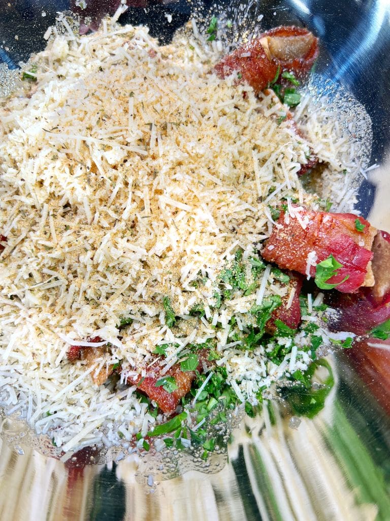 A bowl filled with grated cheese, breadcrumbs, fresh green parsley, and chunks of prosciutto. The ingredients create a colorful and textured mixture in a stainless steel bowl, ready for mixing or topping a dish.