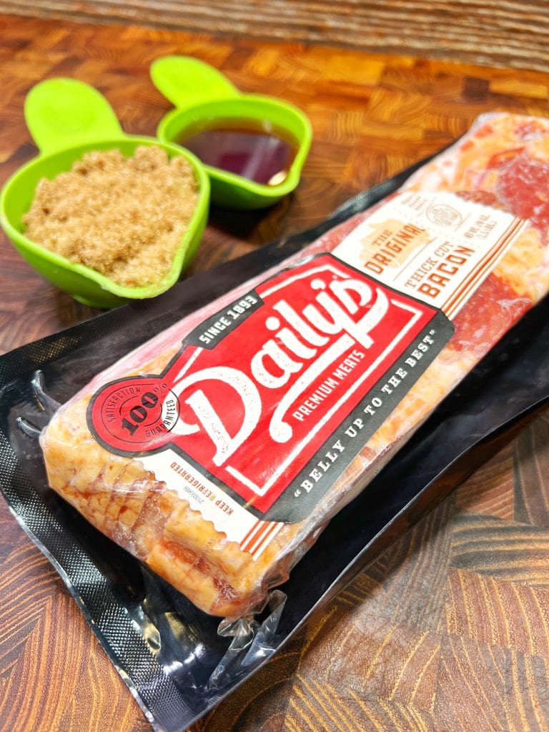 Packaged Dailys premium bacon sits on a wooden surface. Next to it are two green measuring cups, one filled with brown sugar, the other with a dark liquid.