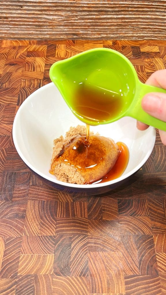 A hand pouring a golden-brown liquid from a green cup onto light brown sugar in a white bowl, set on a wooden textured surface.