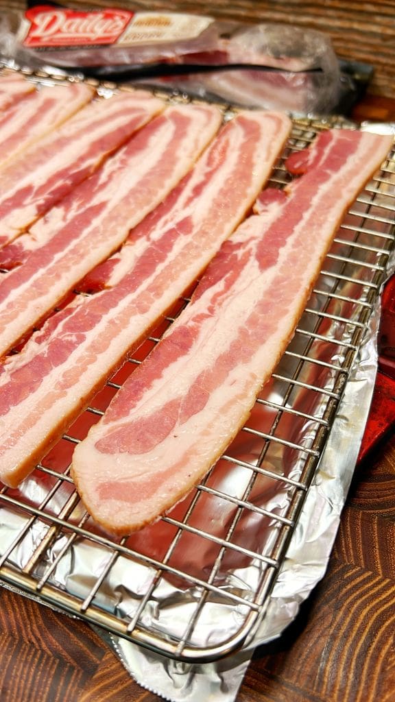 Close-up of several raw bacon strips arranged on a wire rack, ready for cooking. The wire rack is placed over a foil-lined tray, and the bacon showcases its marbled red and white pattern.